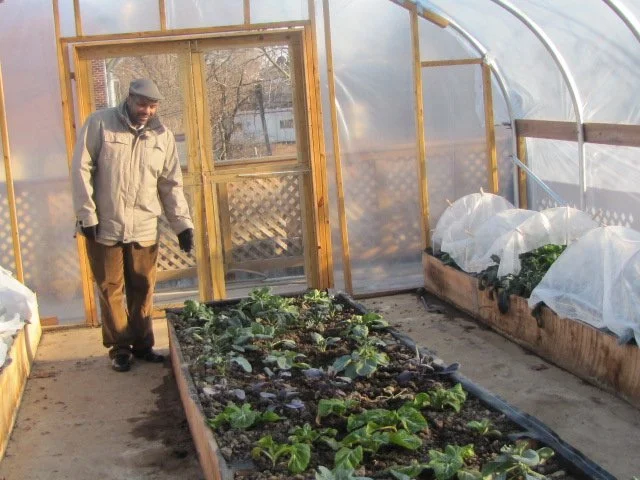 A Black man wearing a tan Kangol cap, khaki coat and brown pants standing inside a greenhouse. He is pointing one hand down to a garden bed full of plants.