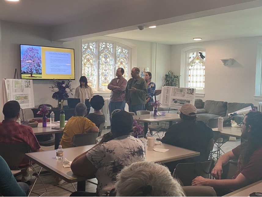 A meeting with several people sitting at tables and several people standing. One standing person is gesturing at a presentation on a TV.
